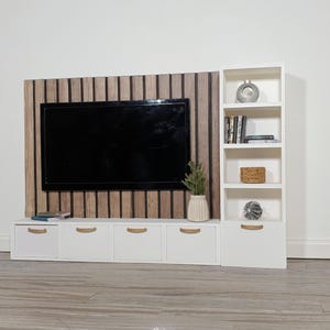 May include: A miniature modern entertainment unit with a black TV, white cabinets, and a wooden accent wall. A white bookshelf is on the right, with decorative items. A small plant sits on the cabinet.