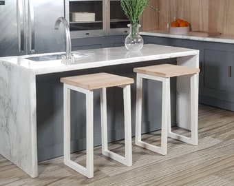 Può includere: Due sgabelli da bar in metallo bianco con sedute in legno. Gli sgabelli sono di fronte a un'isola da cucina in marmo bianco con una base grigia.