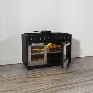 Puede incluir: Un horno negro en miniatura con una puerta de acero inoxidable y una puerta de horno de vidrio. La puerta del horno está abierta, revelando una cesta de comida en el interior. Una sartén está sobre la estufa. El horno está sobre un suelo de madera.