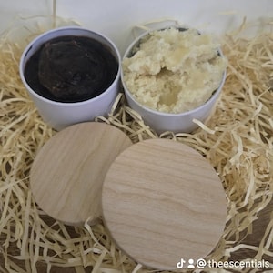 Könnte beinhalten: Zwei weiße zylindrische Behälter mit Kosmetikprodukten, einer gefüllt mit einer dunkelbraunen Substanz und der andere mit einer hellbeigen, strukturierten Creme. Zwei runde, helle Holzdeckel liegen vor den Behältern.