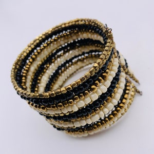 May include: A multi-strand beaded bracelet with black, white, and gold beads. The bracelet is wrapped around itself to create a thick, layered look.