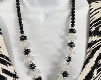 May include: A black and white beaded necklace with a variety of shapes and sizes. The necklace features small black beads, silver-toned accents, and clear, flat, circular beads. The necklace is displayed on a white mannequin.