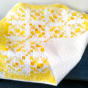 May include: A square quilt featuring a geometric patchwork pattern in shades of bright yellow, golden yellow, and white. The quilt has a solid white backing that is partially folded over to reveal the quilted texture and the vibrant yellow border.