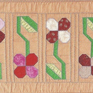 May include: A rectangular quilted table runner in a tan fabric featuring a repeating pattern of appliqued flowers. Each flower has four petals in various shades of red, pink, and white, with green stems and leaves stitched onto the tan background.