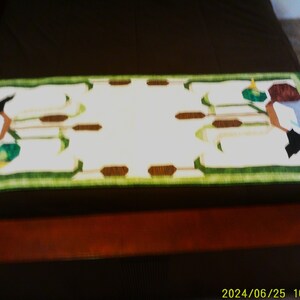 May include: Rectangular decorative table runner with a white center and a green border. The runner features a repeating pattern of stylized figures in brown, green, blue, and black. The runner is placed on a dark brown surface.
