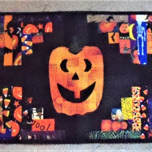 May include: A rectangular fabric table runner featuring a black background with three large orange jack-o'-lanterns. The design includes patchwork accents of Halloween-themed patterns, such as candy corn, skeletons, and pumpkins, arranged in a decorative border.