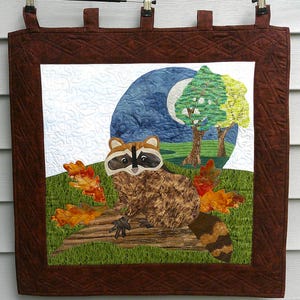 May include: A square fabric wall hanging featuring a quilted scene of a raccoon sitting on a log. The raccoon is brown and black, surrounded by orange and yellow autumn leaves on green grass. A blue moon and two trees are in the background.