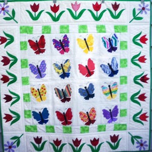 May include: A white quilt with a green and white checkered pattern in the center. The quilt is decorated with a border of red tulips and green leaves. There are 16 colorful butterflies in the center of the quilt.