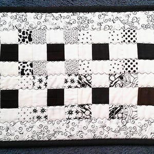 May include: A rectangular, black and white quilted table runner. The design features a checkerboard pattern of solid black squares alternating with white squares, each with a unique black and white pattern. The edges are finished with a black border.