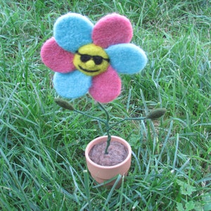 May include: A felt flower with a smiley face and sunglasses in a brown pot. The flower is pink, blue, and yellow.