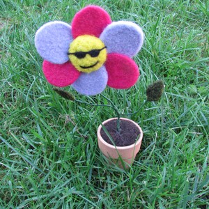 May include: A felt flower with a smiley face wearing sunglasses in a terracotta pot. The flower is pink, purple, and yellow. The pot is sitting on green grass.