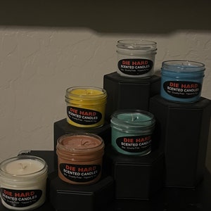 May include: Six scented candles in glass jars with black labels that say "Die Hard Scented Candles" in red letters. The candles are arranged in a pyramid shape on a black surface. The candles are in the colors white, yellow, brown, blue, and green.