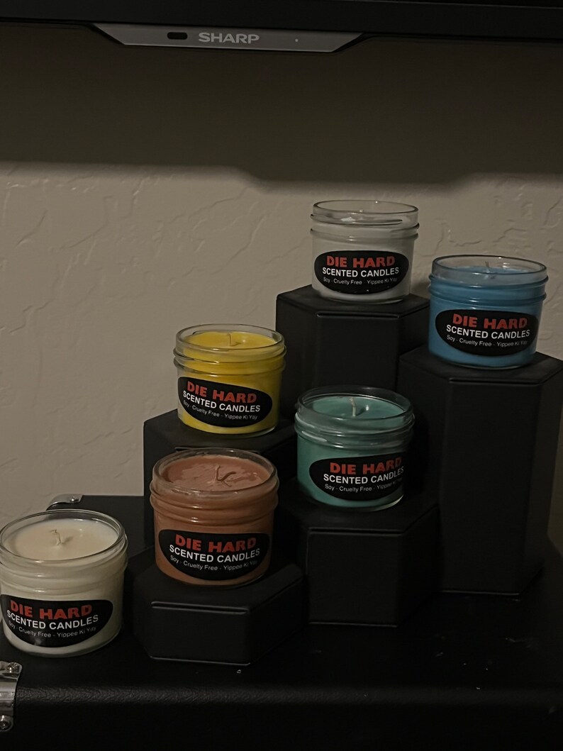 May include: Six scented candles in glass jars with black labels that say "Die Hard Scented Candles" in red letters. The candles are arranged in a pyramid shape on a black surface. The candles are in the colors white, yellow, brown, blue, and green.