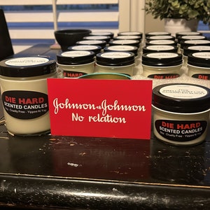May include: A table with a collection of small glass jars with black lids. The jars are labeled "Die Hard Scented Candles" and have a red and white label with the text "Soy-Cruelty Free - Yippee Ki Yay". There is a red card with the text "Johnson & Johnson No relation" on the table.