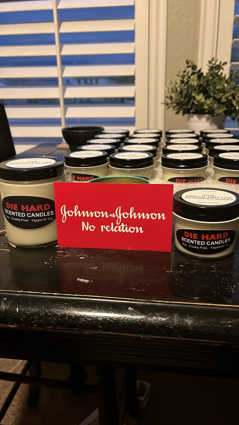 May include: A table with a collection of small glass jars with black lids. The jars are labeled "Die Hard Scented Candles" and have a red and white label with the text "Soy-Cruelty Free - Yippee Ki Yay". There is a red card with the text "Johnson & Johnson No relation" on the table.