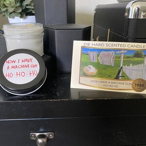 May include: A black and white image of a candle with a label that reads "Now I have a machine gun Ho-Ho-Ho". The candle is on a black wooden surface with a drawer. A card with the text "Die Hard Scented Candles" is visible in the background.