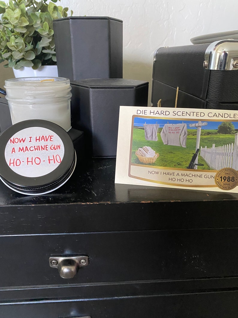 May include: A black and white image of a candle with a label that reads "Now I have a machine gun Ho-Ho-Ho". The candle is on a black wooden surface with a drawer. A card with the text "Die Hard Scented Candles" is visible in the background.