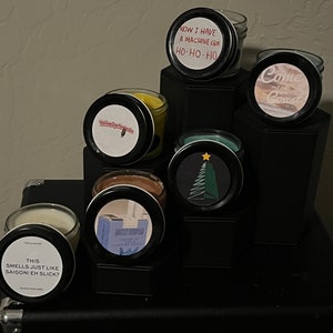 May include: A collection of six candles with various scents and labels. The labels include "Yellow Dye Napkins", "Come out to the Coast", "This smells just like Saigon! Eh Slick?", "Now I have a machine gun Ho-Ho-Ho", "Mister European", and a Christmas tree design.