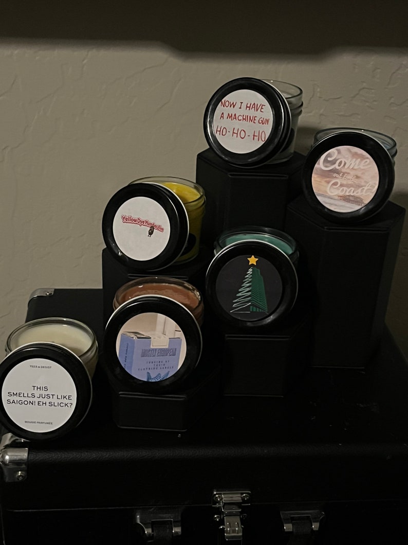 May include: A collection of six candles with various scents and labels. The labels include "Yellow Dye Napkins", "Come out to the Coast", "This smells just like Saigon! Eh Slick?", "Now I have a machine gun Ho-Ho-Ho", "Mister European", and a Christmas tree design.