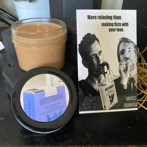 May include: A brown candle in a clear glass jar with a white wick. The candle is sitting on a black surface. A white card with black text reads "More relaxing than making fists with your toes." The card also features a black and white image of two men. The card is next to a black lid with a blue and white label that reads "Mostly European Judging by their clothing labels".
