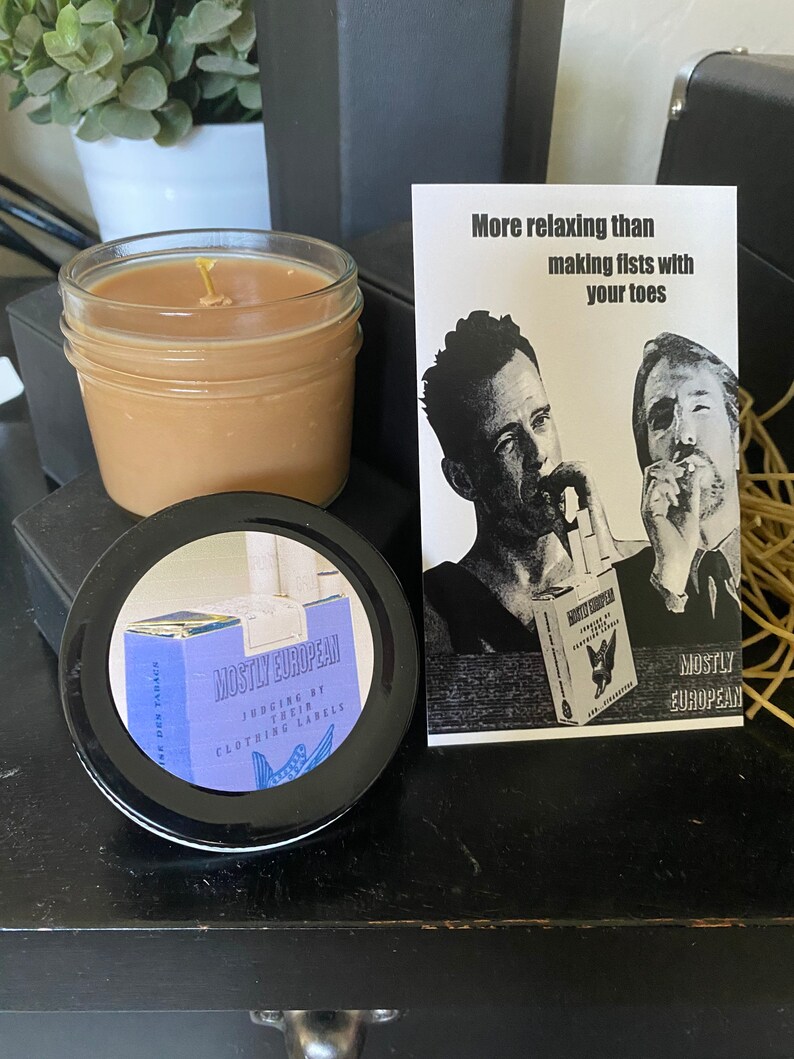 May include: A brown candle in a clear glass jar with a white wick. The candle is sitting on a black surface. A white card with black text reads "More relaxing than making fists with your toes." The card also features a black and white image of two men. The card is next to a black lid with a blue and white label that reads "Mostly European Judging by their clothing labels".