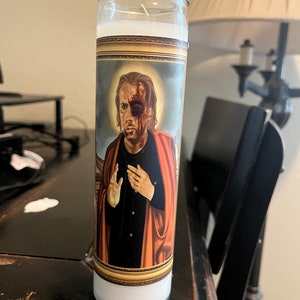 May include: A white candle with a gold-framed image of a man with a black eye and blood on his face. The man is wearing a black shirt and a red robe. The candle is on a dark wooden surface.
