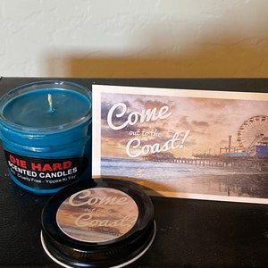 May include: A blue scented candle in a glass jar with a black label that reads "Die Hard Scented Candles Soy-Cruelty Free - Yippee Ki Yay". The candle is next to a postcard with a picture of a beach and a Ferris wheel. The postcard reads "Come out to the Coast!" and has a smaller sticker that reads "Come out to the Coast!" on top of it.