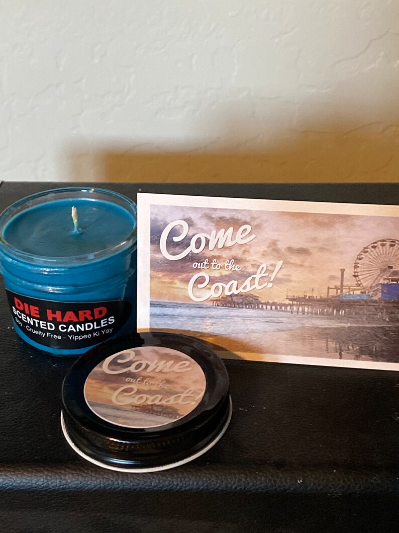 May include: A blue scented candle in a glass jar with a black label that reads "Die Hard Scented Candles Soy-Cruelty Free - Yippee Ki Yay". The candle is next to a postcard with a picture of a beach and a Ferris wheel. The postcard reads "Come out to the Coast!" and has a smaller sticker that reads "Come out to the Coast!" on top of it.