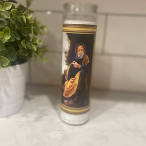 May include: A white candle with a gold-framed image of a man with a beard and a yellow robe. The image is of a religious figure.