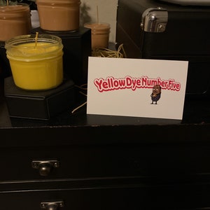 May include: A black wooden dresser with two drawers and a card on top that says "Yellow Dye Number Five" with a cartoon character holding a jar of yellow dye.