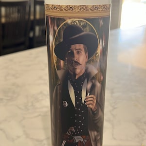 May include: A white candle with a gold-framed image of a man wearing a black hat and coat. The man has a mustache and is holding a cigarette in his right hand.