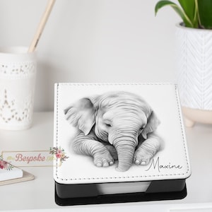 May include: A white and black square notepad holder with a pencil sketch of a baby elephant. The elephant is lying down with its trunk curled. The name "Maxine" is written in cursive on the notepad holder.