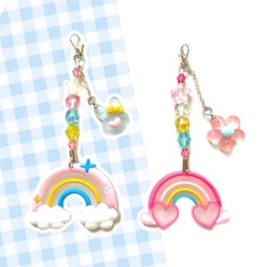May include: Two rainbow charm keychains with colorful beads and charms. One charm features a pink flower and the other a blue heart with a yellow bow. Both charms are attached to a silver chain.