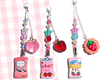 May include: Three pink and white charm keychains with different fruit and candy designs. The charms include a peach, a strawberry, and a strawberry milk carton. Each charm has a silver chain with pink, blue, and white beads and a star charm.
