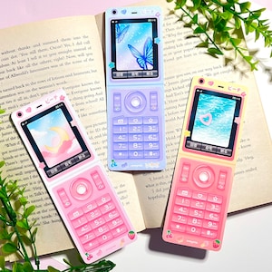 May include: Three colorful flip phones with screens displaying different images. The phones are pink, purple, and yellow, each adorned with decorative stickers of hearts, stars, and fruit. The screens show a moon, butterflies, and a pool.