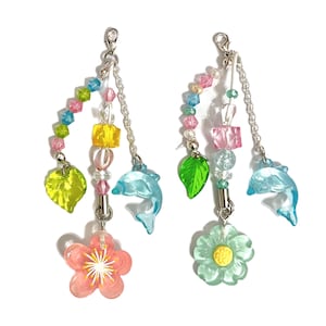 Könnte beinhalten: Zwei bunte Charm-Armbänder mit einer Vielzahl von Perlen, darunter rosa, blau, grün und gelb. Die Charms umfassen einen Delfin, ein Blatt, eine Blume und ein Quadrat. Die Armbänder sind an silbernen Ketten befestigt.