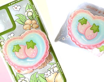 May include: A pink and blue heart-shaped phone grip with two pink strawberries and green leaves. The grip is attached to a clear phone case with a green border and a floral design. The text "good friend" and "pretty good" is visible on the phone case.