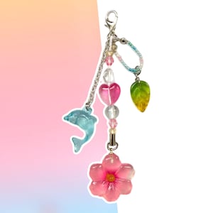 May include: A pink flower charm with a blue dolphin, green leaf, and pink heart charms on a silver chain. The charms are strung with clear and pink beads.