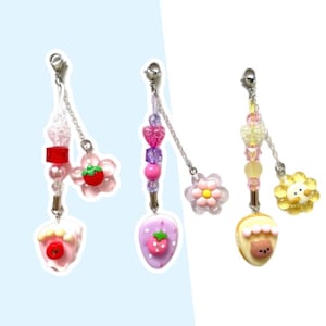 May include: Three colorful beaded phone charms with different charms at the bottom. The first charm is pink and white with a strawberry on top. The second charm is purple and pink with a strawberry on top. The third charm is yellow and white with a bear on top.