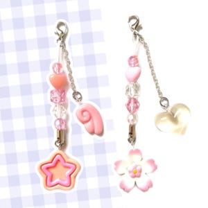 Puede incluir: Dos correas de teléfono con encanto rosa y blanco con cadenas plateadas. Un encanto presenta una estrella rosa, un ala rosa y cuentas rosas y transparentes. El otro encanto presenta una flor blanca, un corazón rosa y cuentas rosas y transparentes.