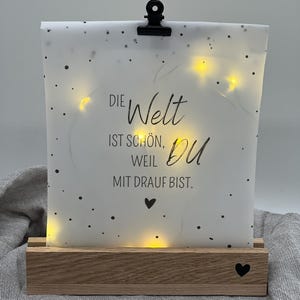 Könnte beinhalten: Leuchtendes Dekoschild mit dem deutschen Text "Die Welt ist schön, weil du mit drauf bist." Das Schild hat einen Holzsockel, eine Papierauflage mit schwarzen Punkten und eine schwarze Klammer. Kleine gelbe Lichter beleuchten das Schild.