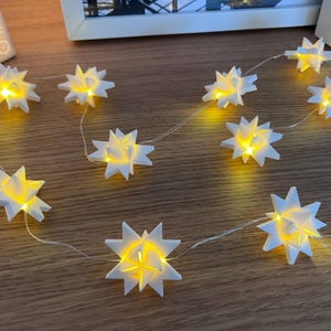 Könnte beinhalten: Eine LED-Lichterkette mit weißen, mehrzackigen sternförmigen Abdeckungen. Jeder Stern strahlt aus seiner Mitte ein warmes gelbes Licht aus. Die Lichter sind durch einen dünnen, transparenten Draht miteinander verbunden.