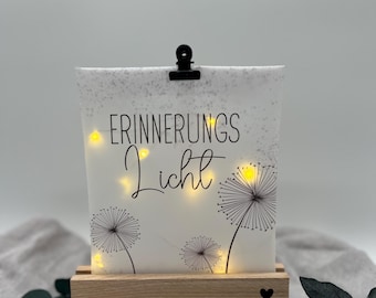 Puede incluir: Una luz decorativa con las palabras "ERINNERUNGS Licht" impresas en una superficie blanca, con ilustraciones de diente de león. La luz se encuentra sobre una base de madera y está iluminada por pequeñas luces amarillas. Un clip negro sujeta la parte superior de la luz.