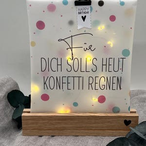 Könnte beinhalten: Dekoratives Leuchtschild mit den Worten "Für Dich soll's heut Konfetti regnen" und "Happy Birthday". Das Schild hat bunte Konfetti-Punkte und wird von einer schwarzen Klammer auf einem Holzsockel gehalten. Ein kleines schwarzes Herz ist auf dem Sockel zu sehen.