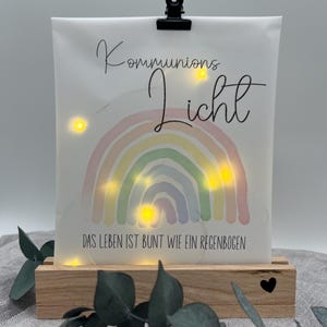 Könnte beinhalten: Ein dekoratives Leuchtschild mit den Worten "Kommunions Licht" und "DAS LEBEN IST BUNT WIE EIN REGENBOGEN" aufgedruckt. Das Schild zeigt ein Regenbogen-Design und wird von einer schwarzen Klammer auf einem Holzsockel gehalten.