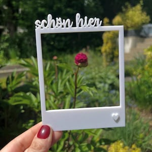 Könnte beinhalten: Ein weißer Rahmen im Polaroid-Stil mit dem deutschen Text "schön hier" oben. Ein kleines Herz befindet sich in der unteren rechten Ecke. Der Rahmen wird vor einer roten Blume in einem Garten gehalten.