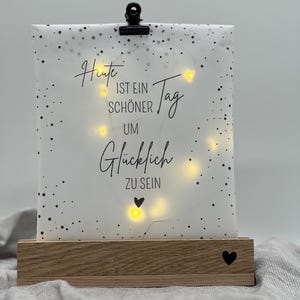 Könnte beinhalten: Ein dekorativer Holzsockel mit einem Herz-Ausschnitt trägt ein weißes Papier mit schwarzen Punkten und dem Text "Heute ist ein schöner Tag um glücklich zu sein." Eine schwarze Klammer hält das Papier, und kleine Lichter beleuchten den Text.