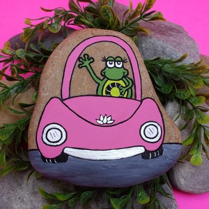 May include: Hand-painted rock art depicting a pink car with a frog driving. The frog is wearing eyelashes and has a green body with a yellow belly. The car has a white flower on the hood and white headlights. The rock is surrounded by green foliage.