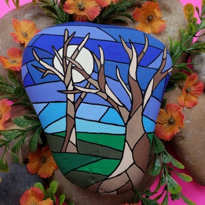 May include: Hand-painted rock art depicting a night scene with two bare trees and a full moon in a blue and purple sky. The rock is painted in a stained glass style with brown, green, blue, and purple colors.