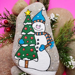 May include: Hand-painted rock art of a snowman holding a Christmas tree. The snowman is wearing a blue hat and scarf with orange stripes. The tree is green with red ornaments and white snow.